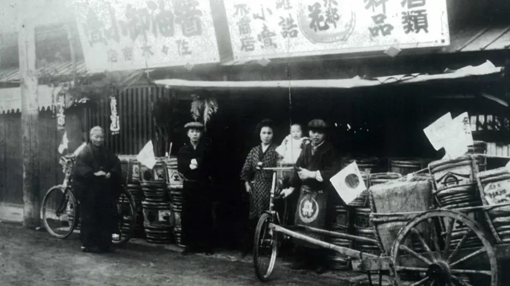 Fotografía histórica en blanco y negro de la fachada de la tienda Sasaki Shouten en 1922 con sus fundadores y carros de reparto cargados de barricas.